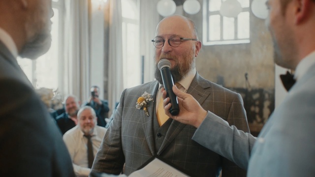 Video Reference N1: Event, Businessperson, Person, Man, Indoor, Standing, Looking, Front, Wearing, Camera, Glasses, Mirror, Suit, Holding, Talking, Hair, People, Wine, Room, Shirt, Glass, Teeth, Phone, Brushing, Human face, Clothing, Ceremony, Microphone, Tie, Wedding, Marriage, Crowd