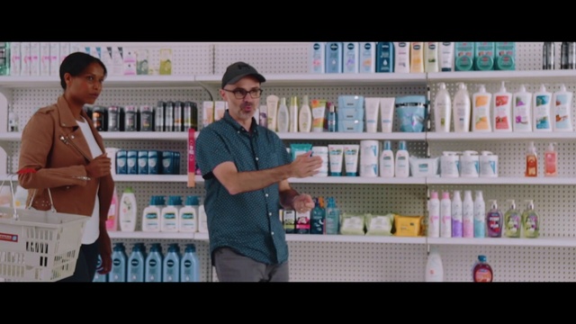 Video Reference N4: Product, Library, Snapshot, Retail, Service, Building, Pharmacy, Customer, Shelf, Screenshot, Person, Indoor, Man, Holding, Table, Sitting, Front, Woman, Standing, Counter, Young, White, Baby, Room, People, Text, Soft drink, Bottle, Convenience store