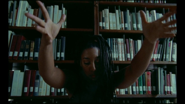 Video Reference N7: Library, Hand, Finger, Snapshot, Arm, Fun, Book, Bookcase, Shelving, Furniture, Person, Indoor, Shelf, Woman, Table, Front, Looking, Holding, Man, Girl, Room, Sitting, Using, Young, Smiling, Head, Shirt, Remote, Blurry, Standing, White, Computer, Red, Video, Text, Human face
