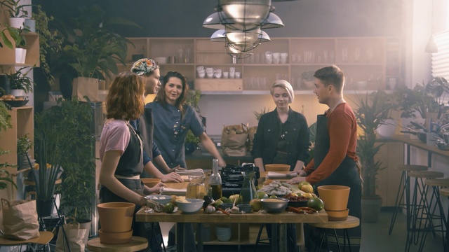 Video Reference N0: Conversation, Meal, Person, Indoor, Table, Woman, Kitchen, Window, Food, People, Dining, Sitting, Restaurant, Man, Standing, Cooking, Eating, Black, Counter, Young, Group, Wine, Plate, Stove, Clothing, Bottle, Smile, Human face, Tableware, Drink, Dining table