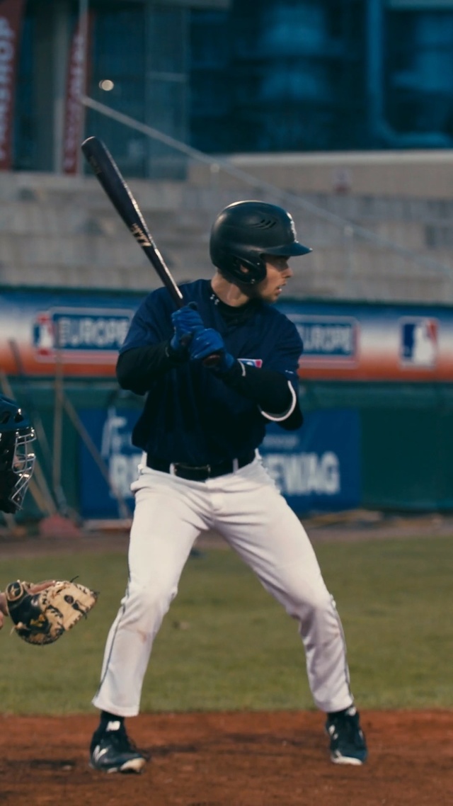 Video Reference N1: Player, Baseball park, Baseball player, Baseball equipment, Sport venue, Sports, Baseball bat, Baseball uniform, Baseball field, Sports equipment, Baseball, Person, Grass, Outdoor, Bat, Game, Building, Swinging, Ball, Field, Holding, Batter, Ready, Pitch, Catcher, Uniform, Standing, Preparing, Watching, Plate, Crowd, Man, Home, Base, Playing, Young, Umpire, Sports uniform, Baseball glove, Athletic game, Glove, Sport, Bat-and-ball games, College baseball, Baseball positions