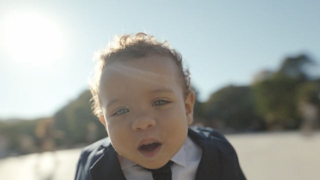 Video Reference N1: Face, Child, Hair, Facial expression, People, Head, Nose, Cheek, Forehead, Male, Person, Outdoor, Young, Looking, Wearing, Woman, White, Boy, Front, Man, Sitting, Shirt, Suit, Smiling, Standing, Eating, Holding, Sky, Toddler, Baby, Human face, Clothing