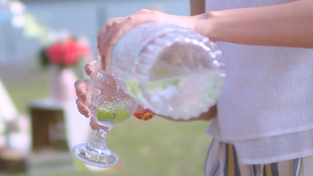 Video Reference N0: Drink, Hand, Glass, Food, Lemonade, Lime, Person, Table, Cup, Cake, Sitting, Small, Holding, Woman, Plate, Piece, Flower, Water, White, Wine, Vase, Eating, Pink, Man, Clear, Sandwich, People, Standing, Bottle, Soft drink
