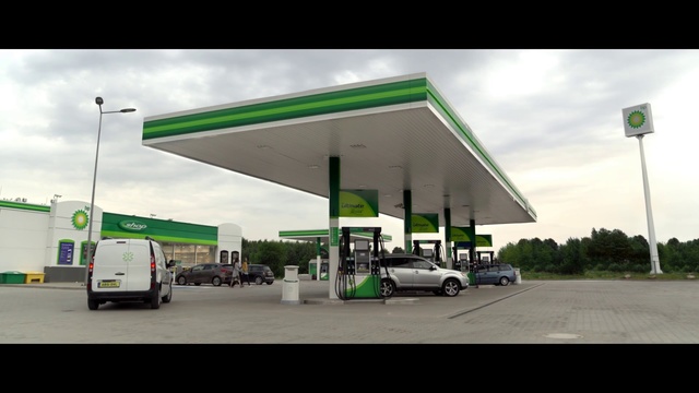 Video Reference N1: Filling station, Gasoline, Fuel, Building, Petroleum, Vehicle, Business, Shade, Car, Gas, Outdoor, Road, Green, Sign, Street, Parked, Runway, Tarmac, Truck, Man, Airplane, Sitting, Plane, Standing, Woman, Large, White, Station, Cloudy, Jet, Driving, People, Traffic, Riding, City, Bus, Air, Group, Sky, Text, Land vehicle, Wheel, Day, Highway