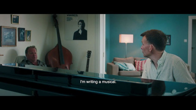 Video Reference N2: Screenshot, Conversation, Pianist, Sitting, Music, Media, Person, Indoor, Man, Table, Laptop, Computer, Front, Looking, Holding, Room, Screen, Black, Desk, Young, Large, Bed, Video, Blue, Wall, Piano, Text, Musical instrument