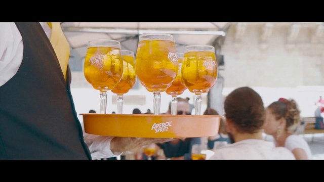 Video Reference N1: Drink, Orange juice, Spritz, Alcoholic beverage, Yellow, Juice, Agua de valencia, Mimosa, Cocktail, Distilled beverage, Person, Indoor, Food, Table, Man, Sitting, Woman, Glass, People, Standing, Orange, Donut, Holding, Restaurant, Display, Cake, Group, Plate, White, Soft drink, Wine glass