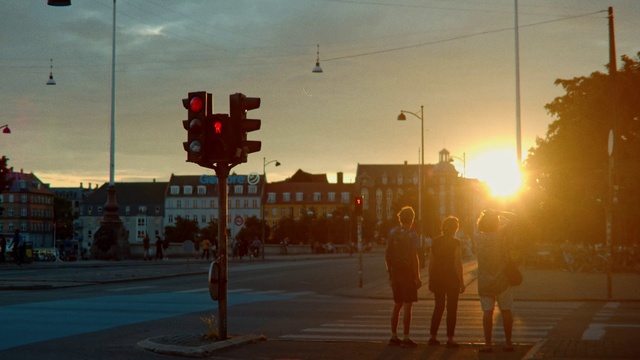 Video Reference N4: Traffic light, Signaling device, Lighting, Sky, Light fixture, Street light, Evening, Cloud, Tree, Street, Person, Outdoor, Light, Traffic, Road, Red, Intersection, Cross, Sunset, Stop, Sun, Standing, Walking, Stopped, City, Signal, Sign, Text